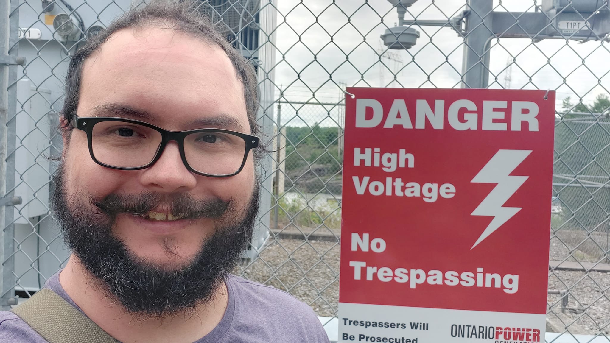 A man short hair and bushy facial hair stands in front of a chain link fence protecting a large transformer, a sign that reads "Danger high voltage, no tresspassing"